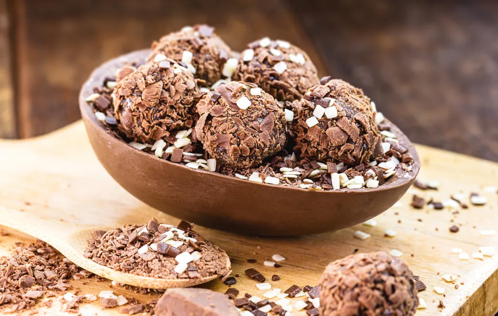 Ovo de páscoa recheado com brigadeiros em cima de uma mesa de madeira próximo a uma colher de pau cheia de farelos de chocolate.