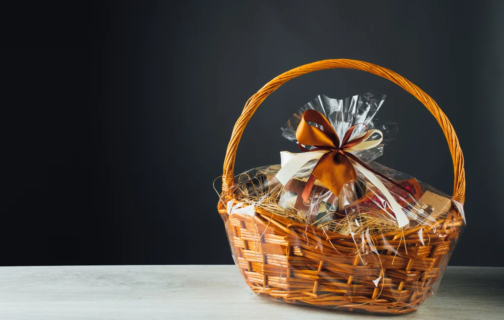 Cesta de presentes para a pascoa: Cesta de vime com alça longa embalada para presente com uma fita dourada fechando a boca do plástivo que envolver a cesta. Cesta com diversos produtos como pães, vinho, queijo e ovo de pascoa. Em uma sala com paredes escuras na cor chumbo e na parte inferior um parte de uma mesa branca na branca que apoia a cesta.