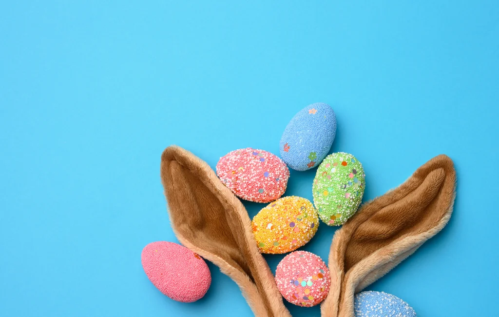 Fundo azul claro com orelhas de coelho marrons surgindo da parte inferior da imagem. Entre as orelhas estão ovos de páscoa coloridos nas cores rosa, verde e amarelo