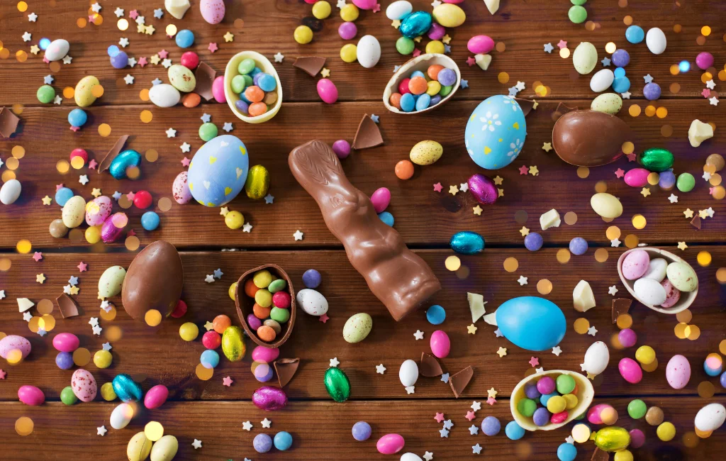 Imagem representativa da páscoa. Fundo da imagem todo em chocolate, em destaque no centro um coelhinho de chocolate e espalhad por todas a imagem, pequenos ovos de pascoa, confeitos como m&ms e algumas amendoas