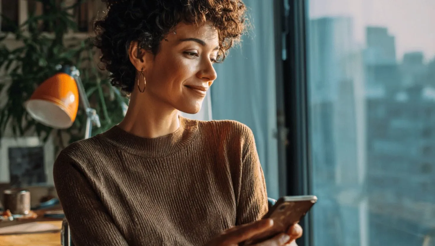 Mulher madura (acima de 40 anos), sorrindo e usando notebook ou smartphone, em ambiente de trabalho ou home office moderno.