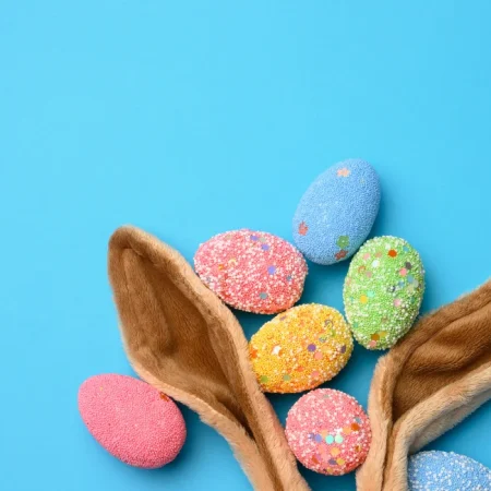 Fundo azul claro com orelhas de coelho marrons surgindo da parte inferior da imagem. Entre as orelhas estão ovos de páscoa coloridos nas cores rosa, verde e amarelo