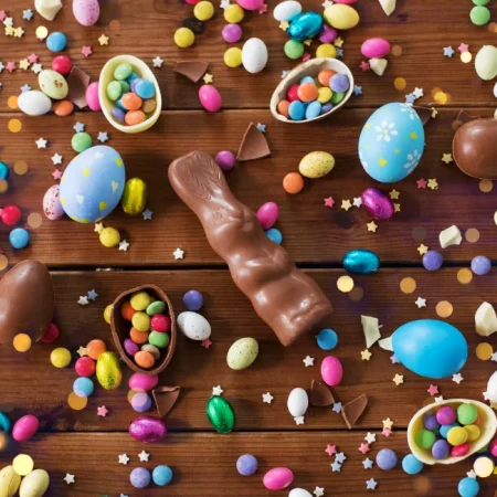 Imagem representativa da páscoa. Fundo da imagem todo em chocolate, em destaque no centro um coelhinho de chocolate e espalhad por todas a imagem, pequenos ovos de pascoa, confeitos como m&ms e algumas amendoas